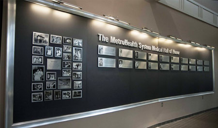 Large, curved wall in wood with a grey laminate and silver frame, made in multiple sections. Metal plate collage printed with historical photos, aluminum lettering, and etched silver plates to honor donors.