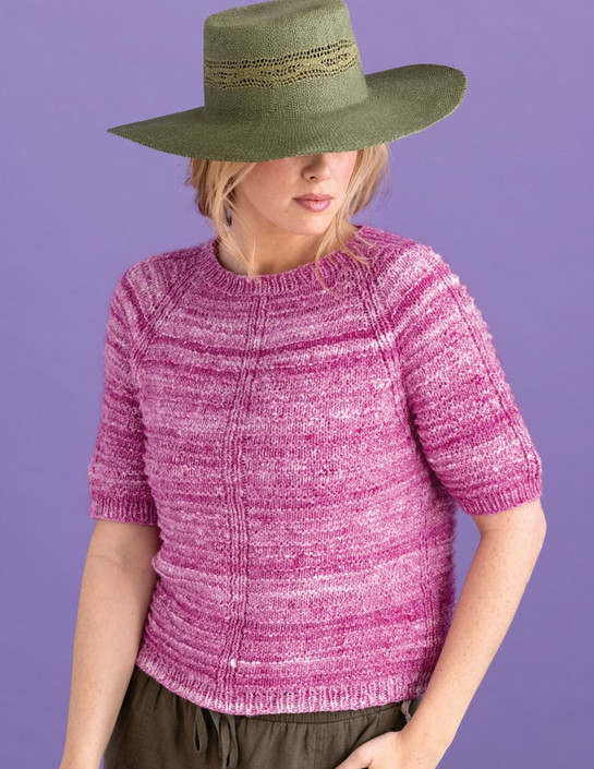 Woman wearing a hand-knit, short-sleeve pink sweater with subtle tonal variations, paired with a wide-brimmed olive green hat against a purple background.