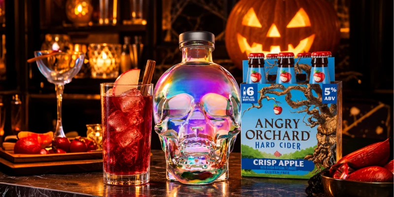 Show a skull-shaped vodka bottle, a red cocktail, and cider on a black bar beside a glowing jack-o’-lantern.