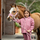 A young rider in a pink outfit holds a brown horse's lead rope, set against a rustic barn backdrop.