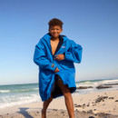 A smiling boy stands on a sandy beach wearing a large blue jacket with the ocean and clear blue sky in the background, conveying a joyful, carefree vibe.