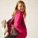 A smiling girl with long blond hair wears a bright pink jacket and carries a backpack with a stuffed bunny and orange water bottle, conveying adventure and joy.