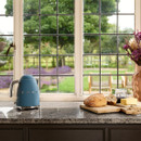 A blue SMEG kettle sits on a granite worktop beside a loaf of bread and butter, with a scenic garden view through the window.