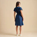 A woman stands with her back to the camera, wearing a denim dress cinched at the waist, complemented by white trainers against a neutral background.