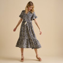 A woman models a flowing, short-sleeved dress with a geometric pattern and tiered hem, styled with brown sandals against a soft beige background.