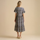 A woman with wavy hair is seen from the back, wearing a patterned, knee-length dress and espadrille sandals against a beige background.