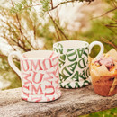 A view of a white mug with MUM written in pink writing and a white mug with dad written in green on a white background
