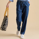 A model walks while wearing loose denim pants, a striped shirt, and white trainers, holding a patterned tote bag.