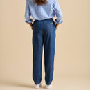 A woman stands facing away, wearing a blue and white striped shirt and loose denim pants, with white trainers on a beige background.