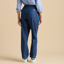A woman in a blue striped shirt and elastic-waist denim jeans turns away, showcasing the back view of her casual outfit against a beige background.