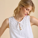 A woman is wearing a white sleeveless top with intricate lace detailing and a tie at the neckline, set against a beige background.