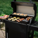A black grill with burgers, sausages, and colorful vegetable skewers cooking, alongside fresh vegetables on a wooden cutting board.
