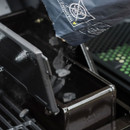 A hand pours charcoal briquettes from a bag into the grill's coal compartment, preparing for outdoor cooking.
