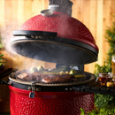 A red Kamado grill with smoke rising, showcasing grilled meats and vegetables, beside a glass of beer and condiments on the table.
