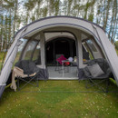 A grey dome tent on a grassy field, open front revealing cosy seating with cushions and blanket, framed by surrounding trees under a blue sky.