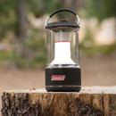 A Coleman lantern sits on a tree stump, emitting a soft glow in a blurred forest background, ideal for camping or outdoor use.