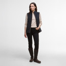 A woman stands confidently in a casual outfit, wearing a navy vest over a light top, black pants, and brown knee-high boots.