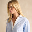 A person wearing a light blue collared shirt with white trim and an embroidered logo, set against a neutral background.