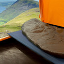 Inside a tent, a dark sleeping bag is set on a camp bed, framed by an open entrance revealing a scenic mountain landscape.