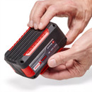 A person holds a compact, red and black battery pack, adjusting its position while revealing its label and textured surface.