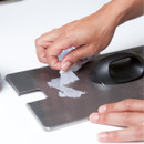 A hand removes a piece of paper from a stainless steel surface, with a cleaning tool visible to the side.