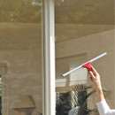 A person uses a red squeegee to clean a large window, revealing a bright, sunny room with plants in the background.