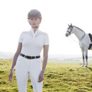 A rider in a white equestrian outfit and helmet stands confidently on a grassy field, with a horse grazing in the background.