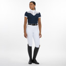 A woman in a navy equestrian shirt, white riding pants, and tall black boots poses confidently against a plain backdrop.