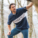 A man in a navy polo shirt with white stripes climbs a tree in a forest, showcasing a casual outdoor style.