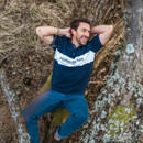 A person relaxes in a natural setting, reclining on a tree while wearing a navy blue polo shirt and jeans, surrounded by grass and woodland.