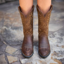 Close-up of a pair of brown cowboy boots with intricate stitching, worn by a person standing on a stone surface.