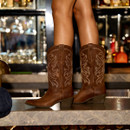 Close-up of brown cowboy boots with embroidered designs, worn by a person standing on a bar counter. Behind, blurred bottles and glasses create a lively bar setting.