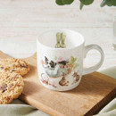 A decorative mug with whimsical animal illustrations sits on a wooden board alongside two chocolate chip cookies.