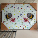 Colourful worktop saver print of two cheerful bees surrounded by flowers and colourful dots, showcased in a protective frame on a marble surface.