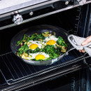 A hand holds a grey towel while removing a skillet with baked eggs, spinach, and zucchini from an oven.