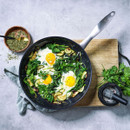 A frying pan with cooked spinach, zucchini, and two sunny-side-up eggs, garnished with parsley, next to a bowl of herbs and mortar.
