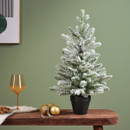 A frosted mini Christmas tree in a pot on a wooden table, accompanied by a glass of wine and golden ornaments, set against a green wall.