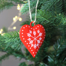 A close up view of a metal red heart-shaped decoration featuring a white snowflake design hanging on a Christmas tree