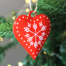 A view of a metal red heart-shaped decoration featuring a white snowflake design hanging on a Christmas tree