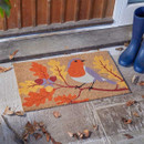A vibrant doormat featuring a colourful robin perched on a branch with autumn leaves, beside blue wellington boots on a stone pathway.