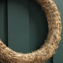 Close-up of a natural straw wreath frame, showing its rough, textured straw surface and fine white thread binding against a dark green door.