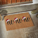 A festive doormat featuring "HO HO HO" in black cursive with Santa hats, placed on a stone floor near a wooden door.