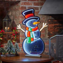 A brightly lit snowman decoration with a red hat and colourful scarf stands against a rustic brick backdrop, exuding a festive charm.