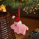 A cute pink pig ornament wearing a red sequined Santa hat hangs from a Christmas tree branch, with twinkling lights in the background.