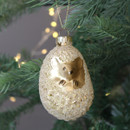 Glittered hedgehog-shaped glass Christmas bauble in a champagne gold finish, featuring a metallic hedgehog face and hanging loop, displayed on a Christmas tree branch with warm fairy lights in the background.