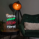 Illuminated Santa please STOP Here! neon sign displayed beside a green armchair and modern floor lamp. Illuminated Santa please STOP Here! neon sign displayed beside a green armchair and modern floor lamp.