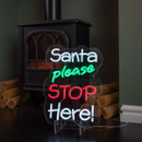 Christmas neon sign with the words Santa please STOP Here! glowing in white, green, and red near a log burner fireplace. Christmas neon sign with the words Santa please STOP Here! glowing in white, green, and red near a log burner fireplace.