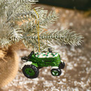 A miniature green tractor ornament decorated with snow and holly, hanging from a snowy pine branch.