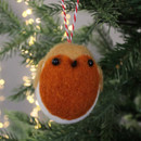 Felt robin ornament with a red breast and black eyes hangs on a Christmas tree branch. Soft lighting and blurred lights create a cozy holiday atmosphere.