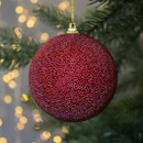 Red textured Christmas ornament hanging on a tree with blurred golden fairy lights in the background, creating a festive and cosy atmosphere.